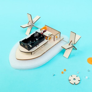 May include: A DIY paddle boat model kit featuring a light-colored wooden frame, a black plastic battery holder, a small electric motor, and two wooden paddle wheels mounted on a metal axle. The base is made of white foam, with small spare parts nearby.