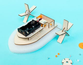May include: A DIY paddle boat model kit featuring a light-colored wooden frame, a black plastic battery holder, a small electric motor, and two wooden paddle wheels mounted on a metal axle. The base is made of white foam, with small spare parts nearby.