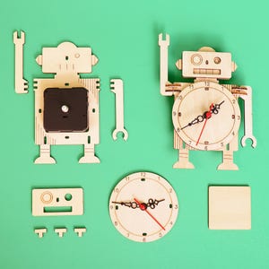 May include: A wooden robot clock kit with a clock face, hands, and a separate clock mechanism. The kit also includes a small wooden square and a rectangular piece with a face.