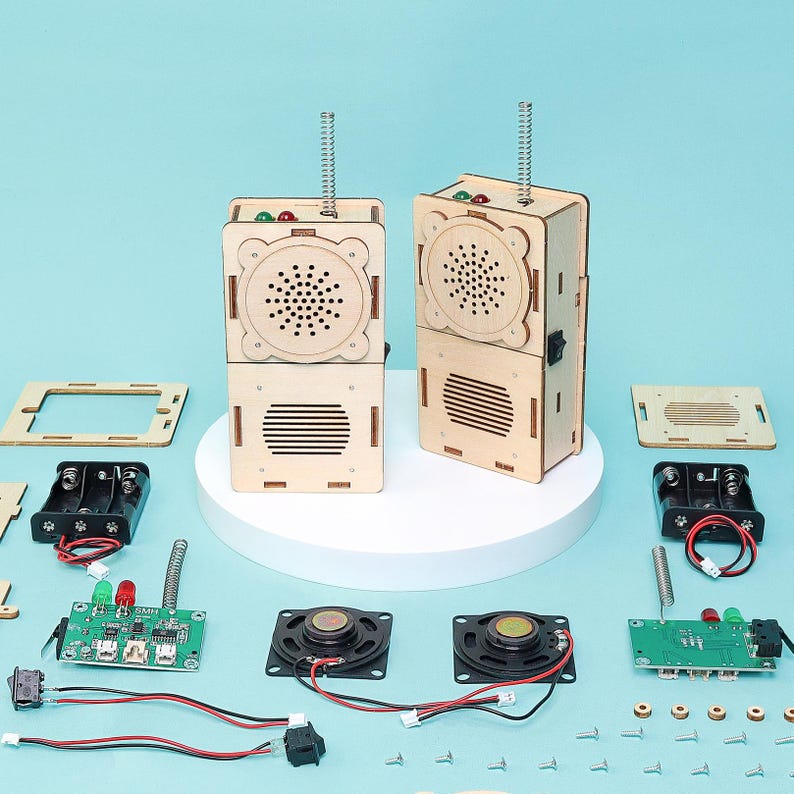 May include: Two assembled wooden radio kits with speakers, antennas, and electronic components. The kits are made of light-colored wood and have a retro design. Various parts, including speakers, circuit boards, and batteries, are arranged around the radios.