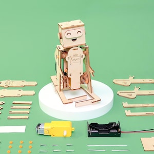May include: A wooden robot kit with a smiling face, assembled and displayed on a white platform. The kit includes various wooden pieces, a yellow motor, a battery holder, and screws, all set against a green background.