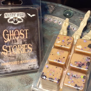 May include: Two packages of soy wax melts. One package has the text "Ghost Stories" and the other has six square wax melts with glitter. The wax melts are tan with purple and black glitter. The text on the package says "Handpoured in Kentucky".