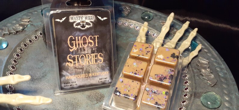May include: Two packages of soy wax melts. One package has the text "Ghost Stories" and the other has six square wax melts with glitter. The wax melts are tan with purple and black glitter. The text on the package says "Handpoured in Kentucky".