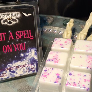 May include: A clear plastic package containing wax melts with pink and purple speckles. Next to it is a package with the text "I PUT A SPELL ON YOU" and the brand name "Mystic Mixxi". The background includes a skeleton hand.