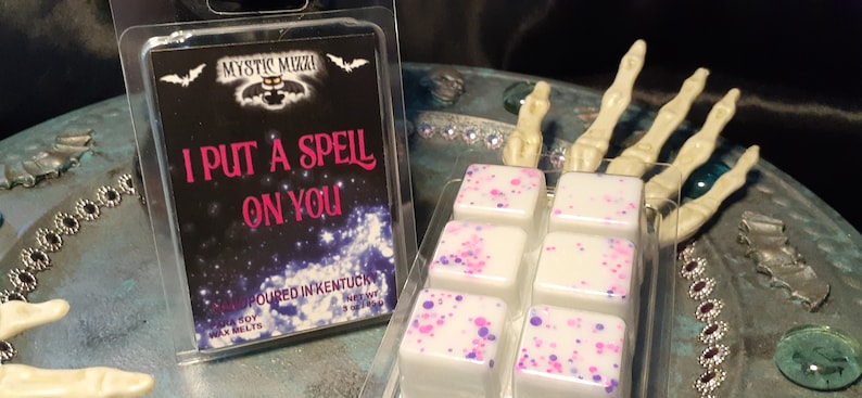 May include: A clear plastic package containing wax melts with pink and purple speckles. Next to it is a package with the text "I PUT A SPELL ON YOU" and the brand name "Mystic Mixxi". The background includes a skeleton hand.