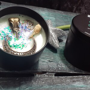 May include: A black candle tin with the text "Witches Cauldron" next to an open candle tin with decorative elements. The open candle has a bronze raven skull, a crescent moon, and glitter. The candle is 2.8 oz.