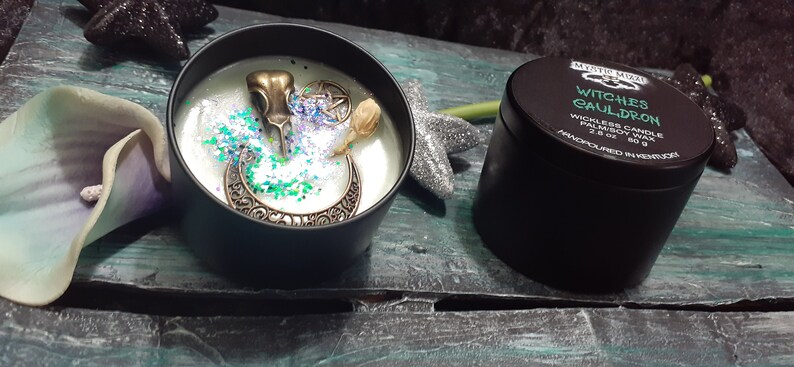 May include: A black candle tin with the text "Witches Cauldron" next to an open candle tin with decorative elements. The open candle has a bronze raven skull, a crescent moon, and glitter. The candle is 2.8 oz.