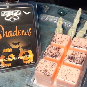 May include: A clear plastic container holds six peach-colored wax melts with dark speckles. A second container displays a black label with the word "Shadows" in orange, along with Halloween-themed graphics. The label reads "Handpoured in Kentucky."