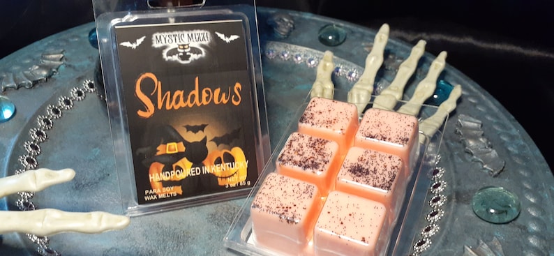 May include: A clear plastic container holds six peach-colored wax melts with dark speckles. A second container displays a black label with the word "Shadows" in orange, along with Halloween-themed graphics. The label reads "Handpoured in Kentucky."