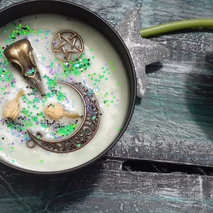 May include: A black candle tin with a light green wax, decorated with a bronze raven skull, crescent moon, and pentagram. The candle is sprinkled with green and purple glitter. A second tin reads "Witches Cauldron" and "5 oz 140 g".