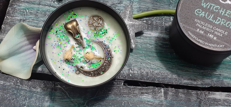 May include: A black candle tin with a light green wax, decorated with a bronze raven skull, crescent moon, and pentagram. The candle is sprinkled with green and purple glitter. A second tin reads "Witches Cauldron" and "5 oz 140 g".