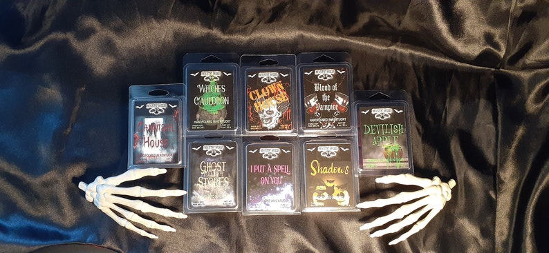 May include: A collection of Halloween-themed wax melts in clear plastic packaging, arranged on a black satin surface. The labels feature spooky graphics and text like "Witches Cauldron" and "Clown House." White plastic skeleton hands frame the display.