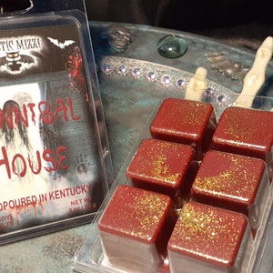 May include: A clear plastic package containing six red wax melts with gold glitter, next to a package with the text "Cannibal House". The packaging has a spooky theme with a ghostly image and the words "Mystic Muzza".