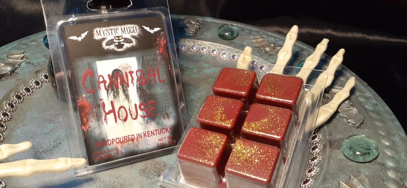 May include: A clear plastic package containing six red wax melts with gold glitter, next to a package with the text "Cannibal House". The packaging has a spooky theme with a ghostly image and the words "Mystic Muzza".