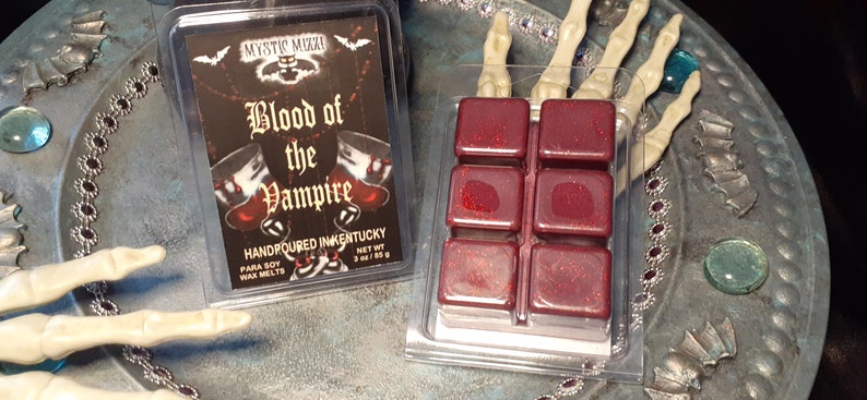 May include: A clear plastic container with six red wax melt cubes and a label that reads "Blood of the Vampire." The label is black with red and white text and features a bat and two goblets. The wax melts are 3 oz / 85 g.