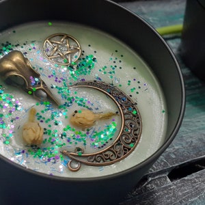 May include: A black candle tin filled with pale green wax, decorated with a bronze raven skull, a crescent moon, a pentagram, and dried flowers. The wax is sprinkled with green and silver glitter. A second black tin is visible in the background.