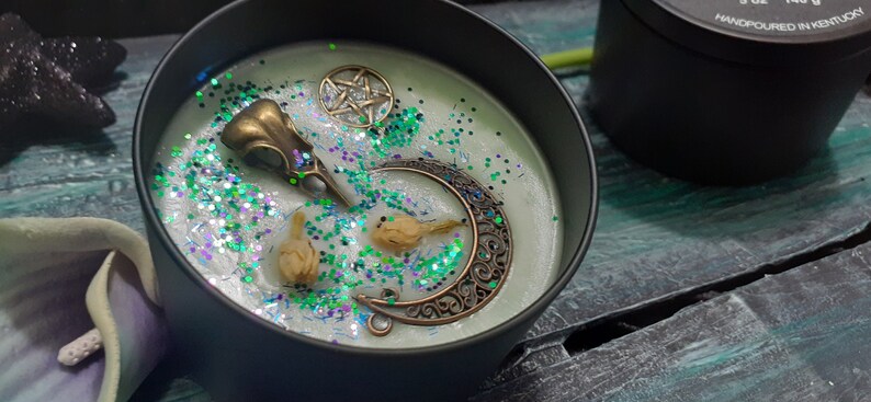 May include: A black candle tin filled with pale green wax, decorated with a bronze raven skull, a crescent moon, a pentagram, and dried flowers. The wax is sprinkled with green and silver glitter. A second black tin is visible in the background.
