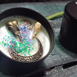 May include: A black candle tin with a decorative design. The candle is decorated with a bronze raven skull, a crescent moon, and a pentagram. The candle is surrounded by green and purple glitter. The tin next to it has the text "Witches Cauldron".