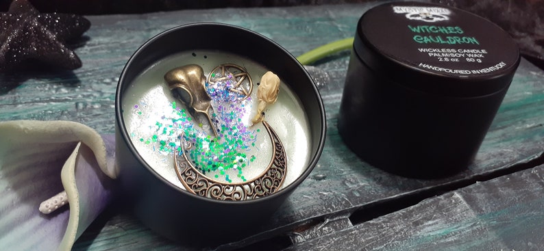 May include: A black candle tin with a decorative design. The candle is decorated with a bronze raven skull, a crescent moon, and a pentagram. The candle is surrounded by green and purple glitter. The tin next to it has the text "Witches Cauldron".
