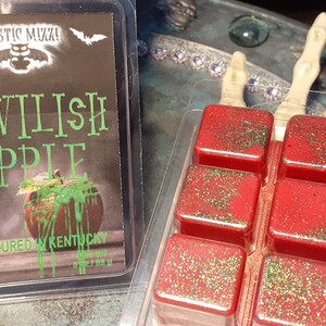 May include: Two containers of wax melts. One has a label that reads "Devilish Apple" with an apple graphic. The other contains six red wax melt cubes with green glitter. The label says "Handpoured in Kentucky".