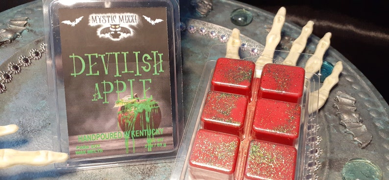 May include: Two containers of wax melts. One has a label that reads "Devilish Apple" with an apple graphic. The other contains six red wax melt cubes with green glitter. The label says "Handpoured in Kentucky".