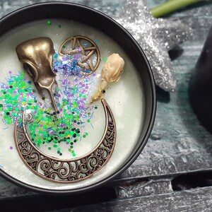 May include: A black candle tin with a light green wax, decorated with bronze charms, glitter, and a dried flower. The charms include a bird skull, a crescent moon, and a pentagram. The label reads "Witches Cauldron".