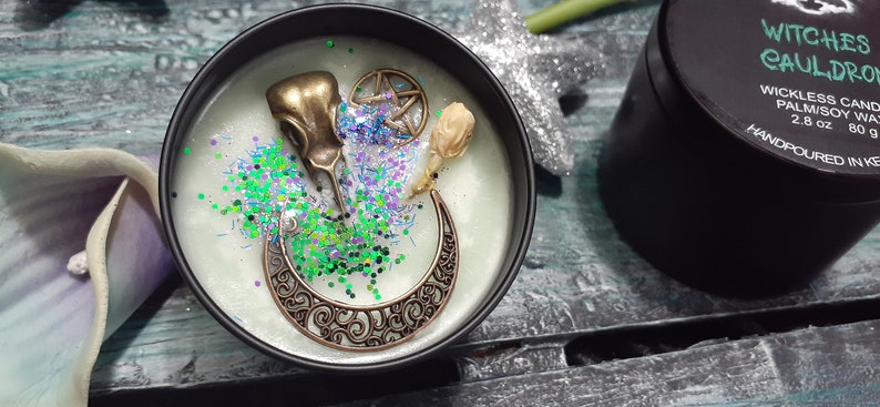 May include: A black candle tin with a light green wax, decorated with bronze charms, glitter, and a dried flower. The charms include a bird skull, a crescent moon, and a pentagram. The label reads "Witches Cauldron".