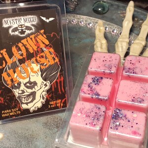 May include: A clear plastic package containing six pink wax melts with purple glitter and a package of wax melts with a scary clown graphic. The text on the package reads "CLOWN HOUSE" and "Mystic Muzzi".