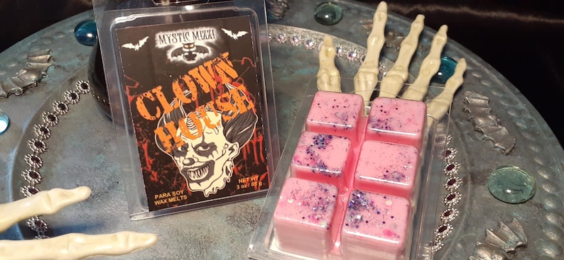 May include: A clear plastic package containing six pink wax melts with purple glitter and a package of wax melts with a scary clown graphic. The text on the package reads "CLOWN HOUSE" and "Mystic Muzzi".