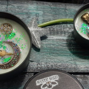 May include: Two black tin candles with pale green wax, decorated with bronze-colored crescent moons, bird skulls, and pentagrams. Green glitter accents the wax. One candle lid is visible, with the text "Witches Cauldron".