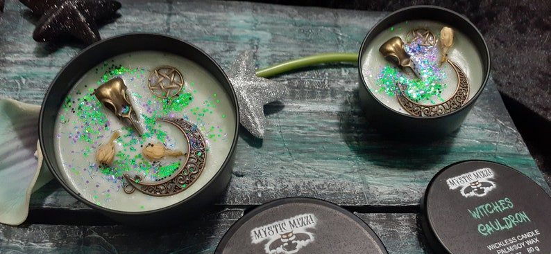 May include: Two black tin candles with pale green wax, decorated with bronze-colored crescent moons, bird skulls, and pentagrams. Green glitter accents the wax. One candle lid is visible, with the text "Witches Cauldron".