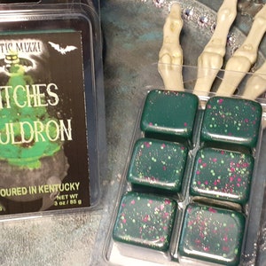 May include: Two packages of wax melts. One package has a label that reads "Witches Cauldron" with a cauldron graphic. The other package contains six dark green wax melt cubes with pink and white speckles. The wax melts are in a clear plastic container.