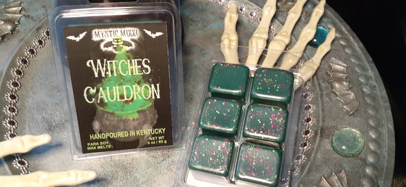 May include: Two packages of wax melts. One package has a label that reads "Witches Cauldron" with a cauldron graphic. The other package contains six dark green wax melt cubes with pink and white speckles. The wax melts are in a clear plastic container.