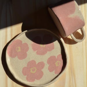 Può includere: Un set di tazza e piatto in ceramica fatti a mano. La tazza e il piatto sono di colore rosa chiaro con un motivo floreale. Il piatto ha uno sfondo beige con fiori rosa. La tazza ha una forma quadrata con un manico.