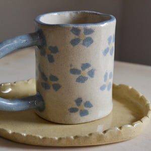 Peut inclure: Tasse en céramique faite à la main avec une anse bleu clair et un motif floral. La tasse est de couleur crème avec des motifs de fleurs bleues et jaunes. Elle repose sur une soucoupe assortie avec un bord festonné. La tasse mesure environ 10 cm de haut.
