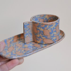 Può includere: Una tazza in ceramica con manico piatto e rettangolare e un piattino ovale abbinato. Entrambi gli oggetti presentano un motivo maculato con forme blu, beige e marrone chiaro su una superficie liscia e smaltata.