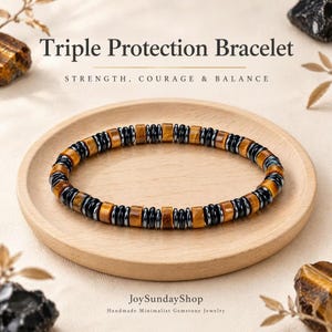 Peut inclure: Un bracelet composé de perles cylindriques en œil de tigre alternées avec des disques plats en hématite noire. Le texte Triple Protection Bracelet, Strength, Courage & Balance et JoySundayShop Handmade Minimalist Gemstone Jewelry est visible.