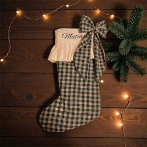 May include: A Christmas stocking with a green and beige check pattern, a cream-coloured top embroidered with the name "Matias" in black, and a matching check bow. The stocking is decorated with a sprig of evergreen and fairy lights.