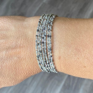 Puede incluir: Un brazalete de múltiples hilos hecho de pequeñas cuentas grises, plateadas y negras.