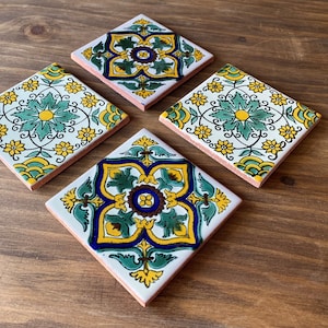 Pode incluir: Quatro bases de cerâmica quadradas com desenhos florais coloridos. Cada base apresenta um fundo branco com padrões amarelos, verdes e azuis. As bordas são de cor rosa claro e as bases estão dispostas sobre uma superfície de madeira.