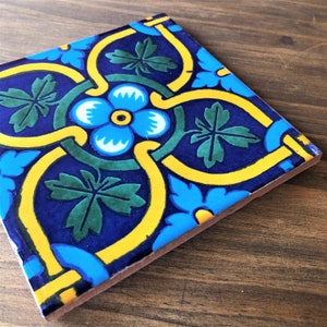Puede incluir: Azulejo cerámico cuadrado con diseño floral. El azulejo presenta un fondo azul oscuro con un patrón floral amarillo, azul claro y verde. El diseño incluye una flor central y detalles de hojas.