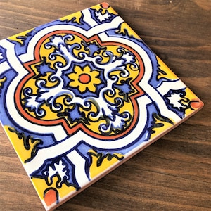 May include: Square ceramic tile with a colorful floral design. The tile features a central yellow flower with an orange center, surrounded by blue and white patterns. The edges are outlined in orange and black.