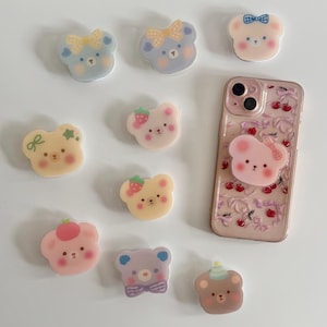 May include: An assortment of colourful phone grips featuring cartoon bears with different accessories. The grips are made of clear plastic with a pink, yellow, blue, or purple bear design. The bears have different accessories, including bows, strawberries, and a party hat.