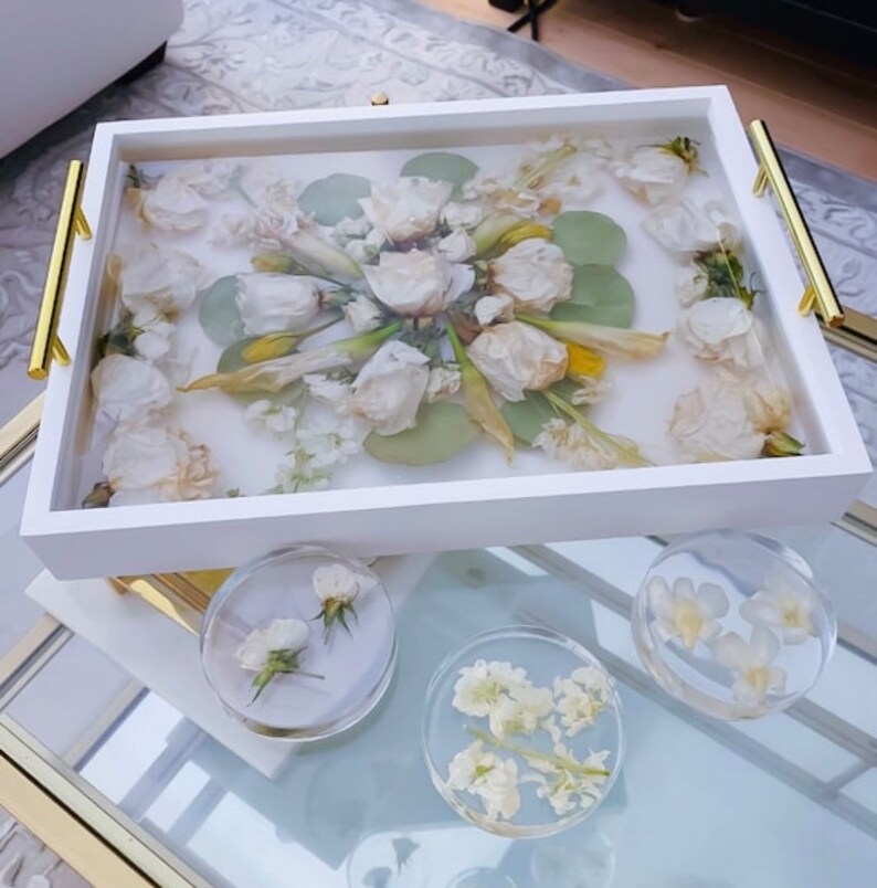 May include: A white tray with gold handles, filled with pressed white flowers and greenery. The tray is on a glass table with three smaller trays filled with pressed flowers.