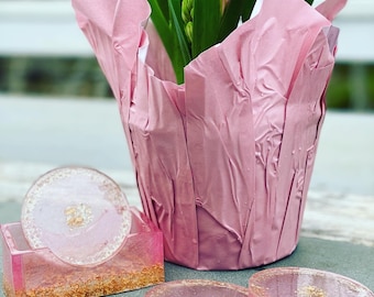 May include: Four pink and gold resin coasters with a pink flower pot wrapped in pink tissue paper in the background.