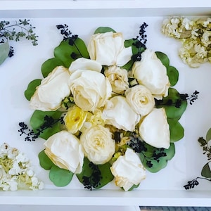 May include: A white tray with a bouquet of dried white roses and green leaves. The roses are arranged in a circular pattern and are surrounded by green leaves. The tray is white and has a gold trim.