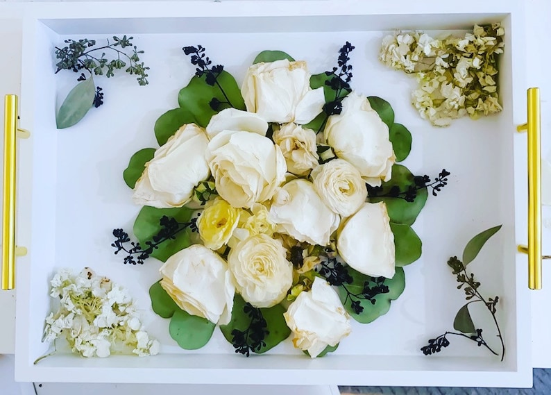 May include: A white tray with a bouquet of dried white roses and green leaves. The roses are arranged in a circular pattern and are surrounded by green leaves. The tray is white and has a gold trim.