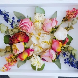 May include: A white tray with a pressed flower arrangement. The arrangement features pink, white, and red roses, white carnations, and blue delphiniums.
