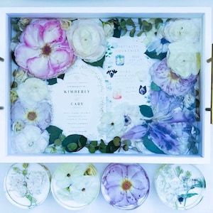 May include: A white tray with a glass top filled with pressed flowers and greenery. The tray also contains a wedding invitation with the names Kimberly and Cary. The tray is surrounded by four smaller white trays with pressed flowers.
