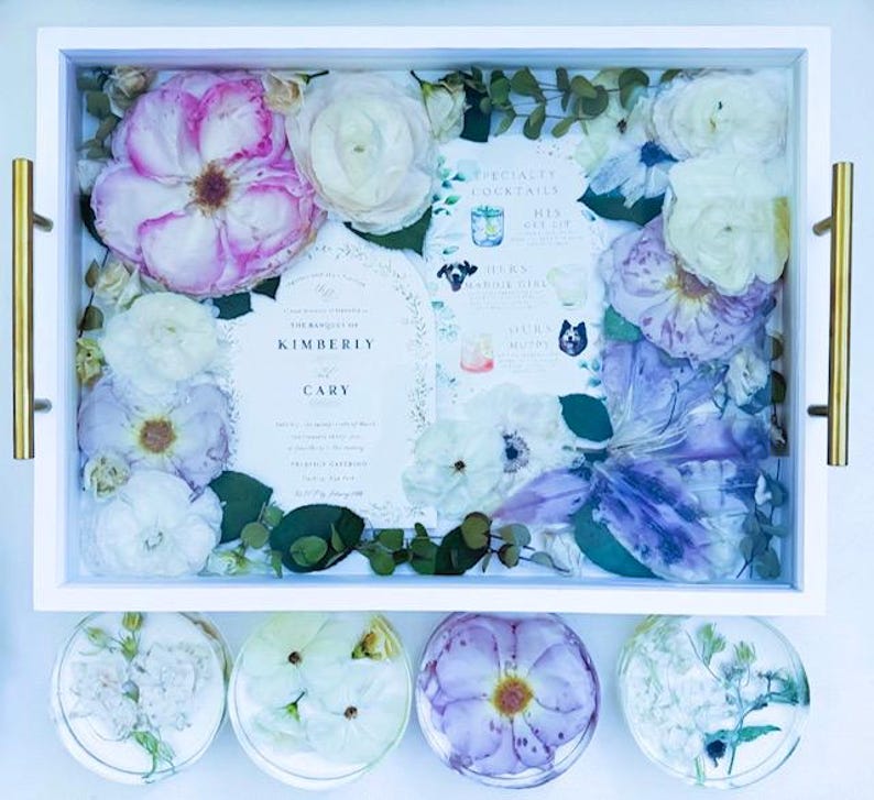 May include: A white tray with a glass top filled with pressed flowers and greenery. The tray also contains a wedding invitation with the names Kimberly and Cary. The tray is surrounded by four smaller white trays with pressed flowers.